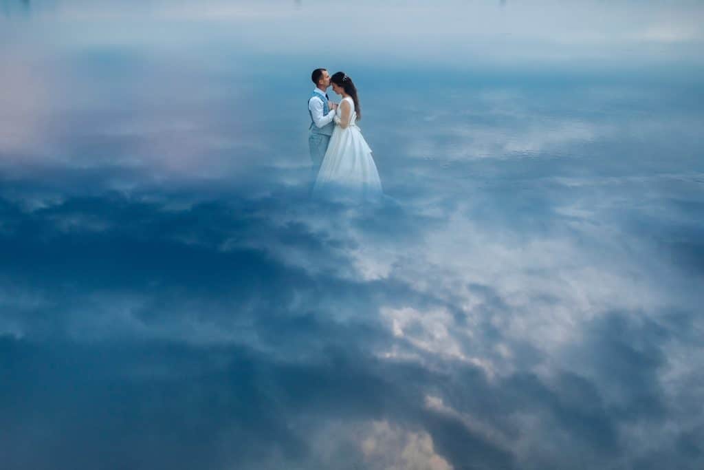 Stock photo of a just married couple in suit and wedding dress standing face to face holding hands. Imitation of standing surrounded by clouds in the sky.