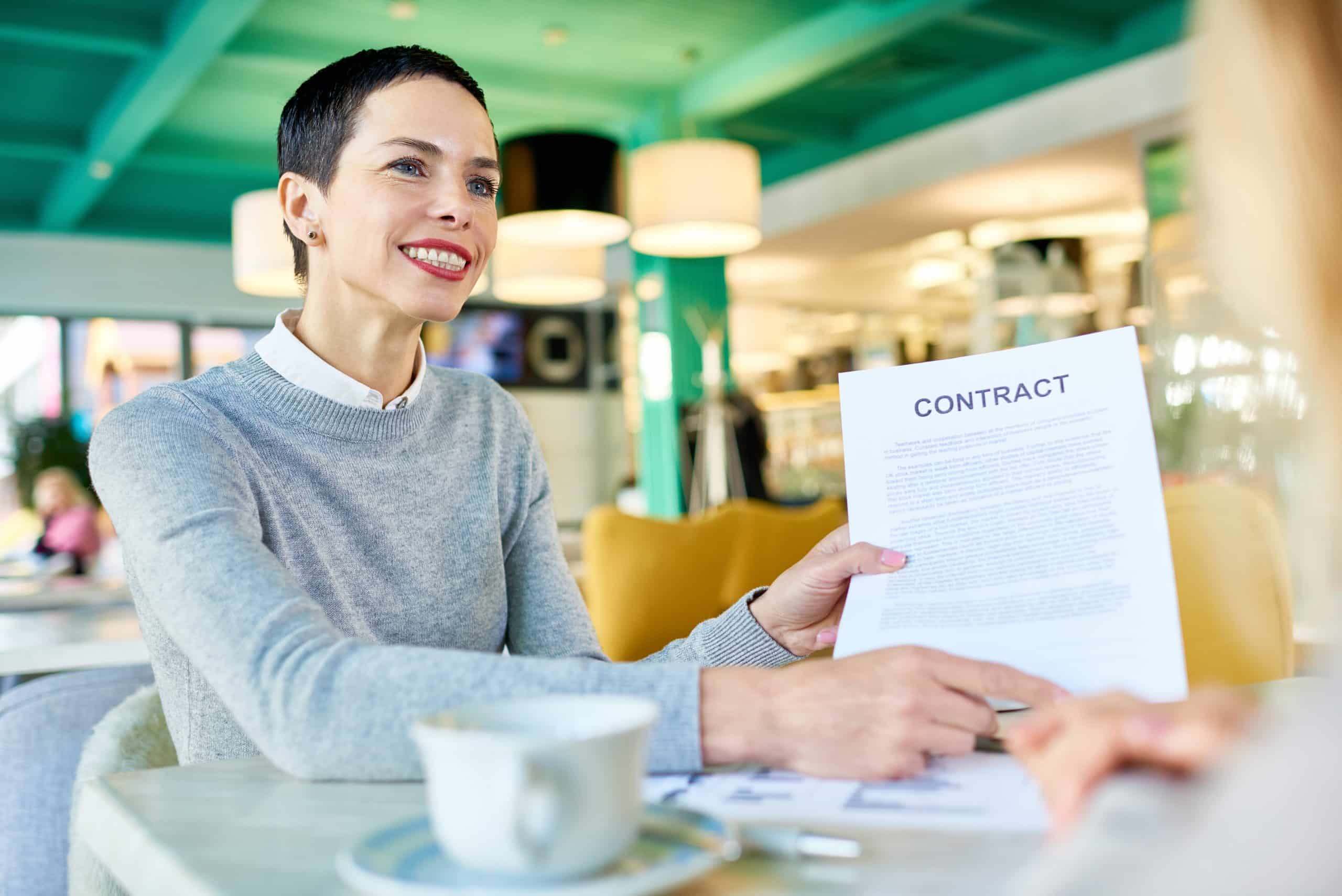 Portrait of powerful businesswoman smiling victoriously handing contract to partner during business meeting in cafe