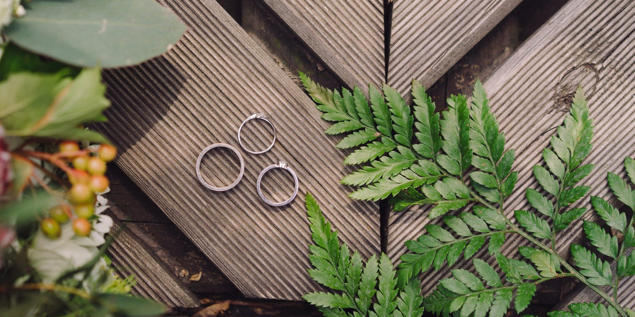 Botanic bridal chic. Bouquet, wedding rings and engagement ring on wooden background with greenery and fern leaves. Copy space, top view, flat lay. Rustic composition.