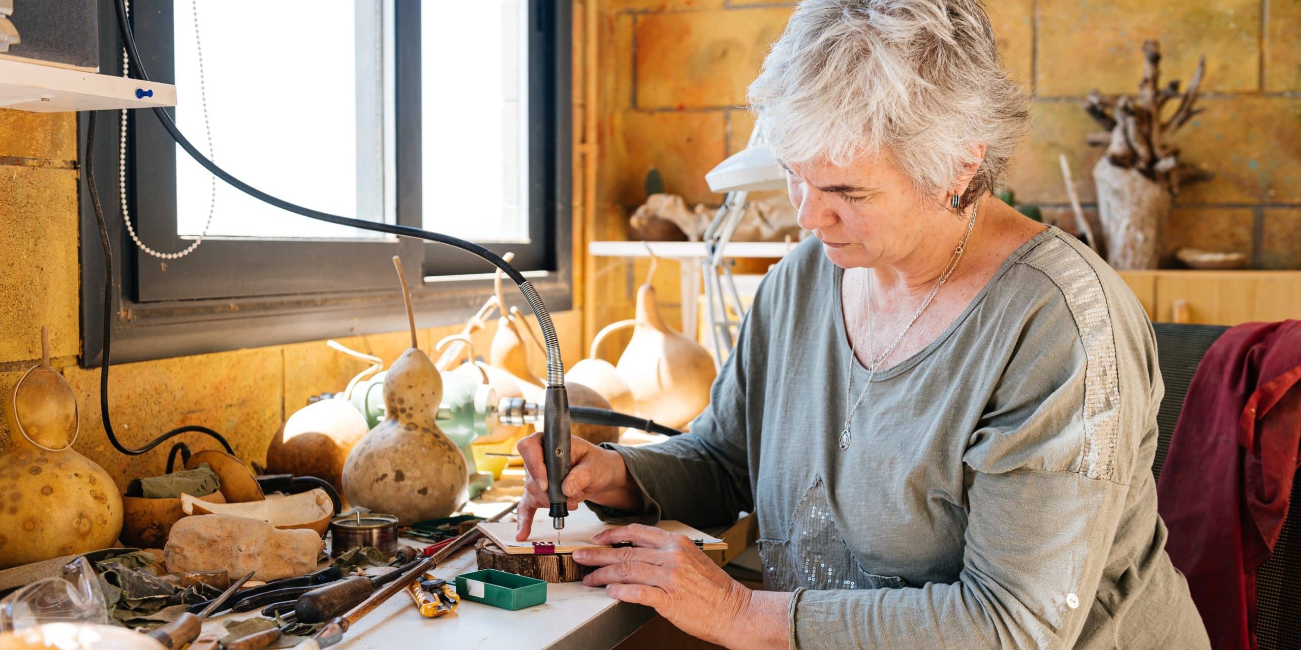 Side view of concentrated elderly female artisan in casual clothes looking down while sitting at table and using engraving pen on hardcover during making of handmade notebook at home