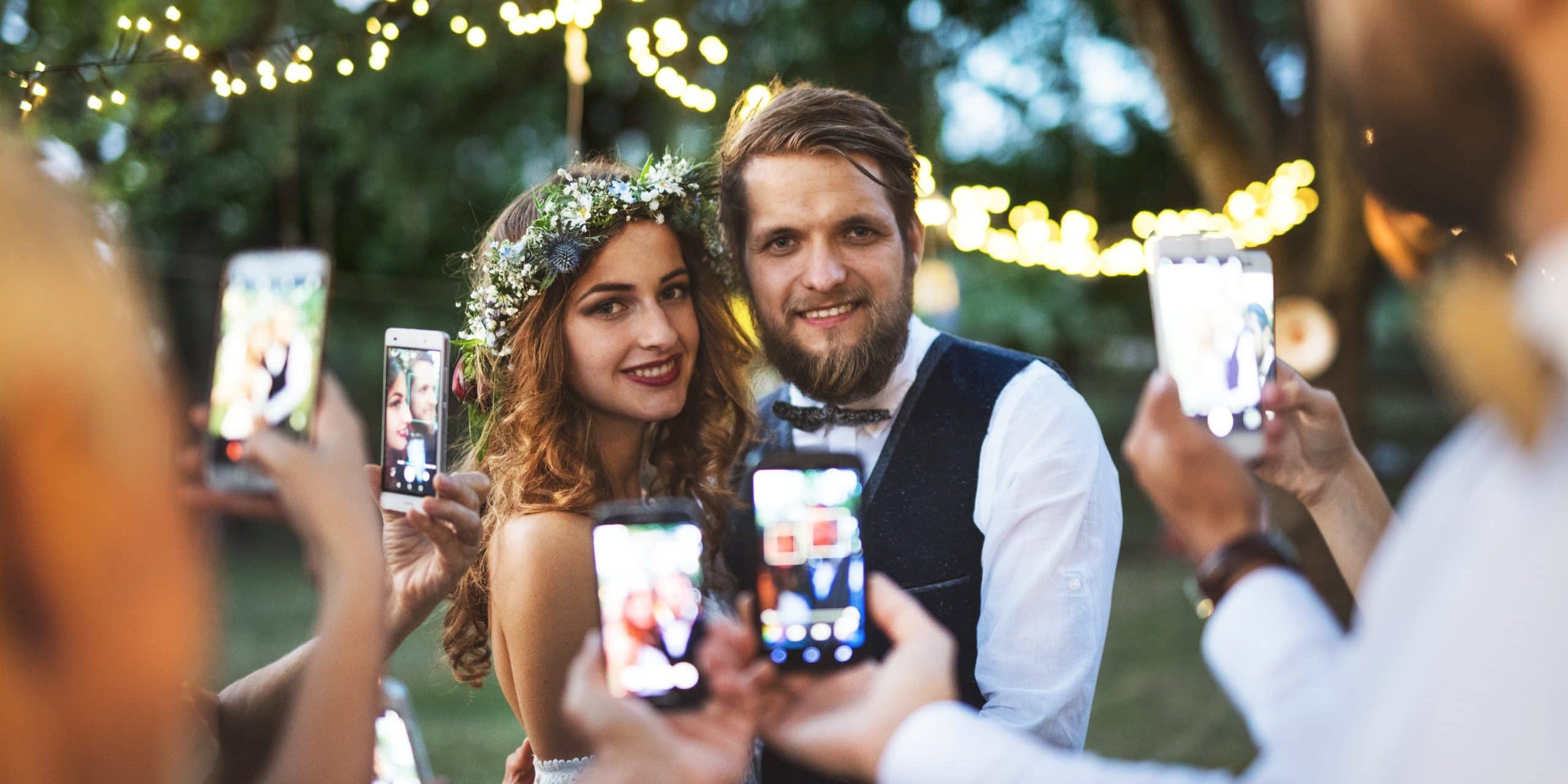 Guests with smartphones taking photo of bride and groom at wedding reception outside in the backyard.