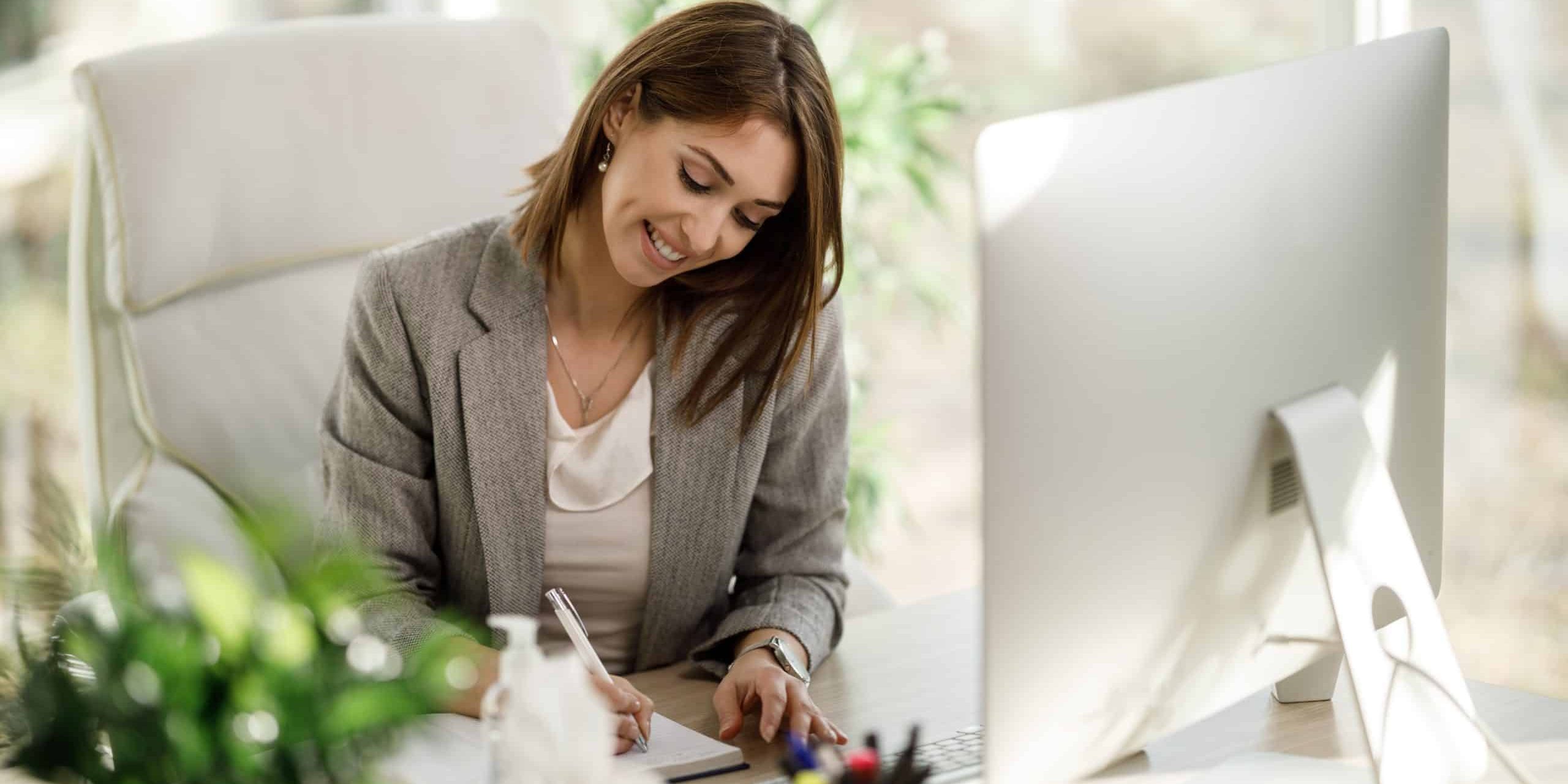 A smiling business woman write something down in a notebook while using a computer in her home office.