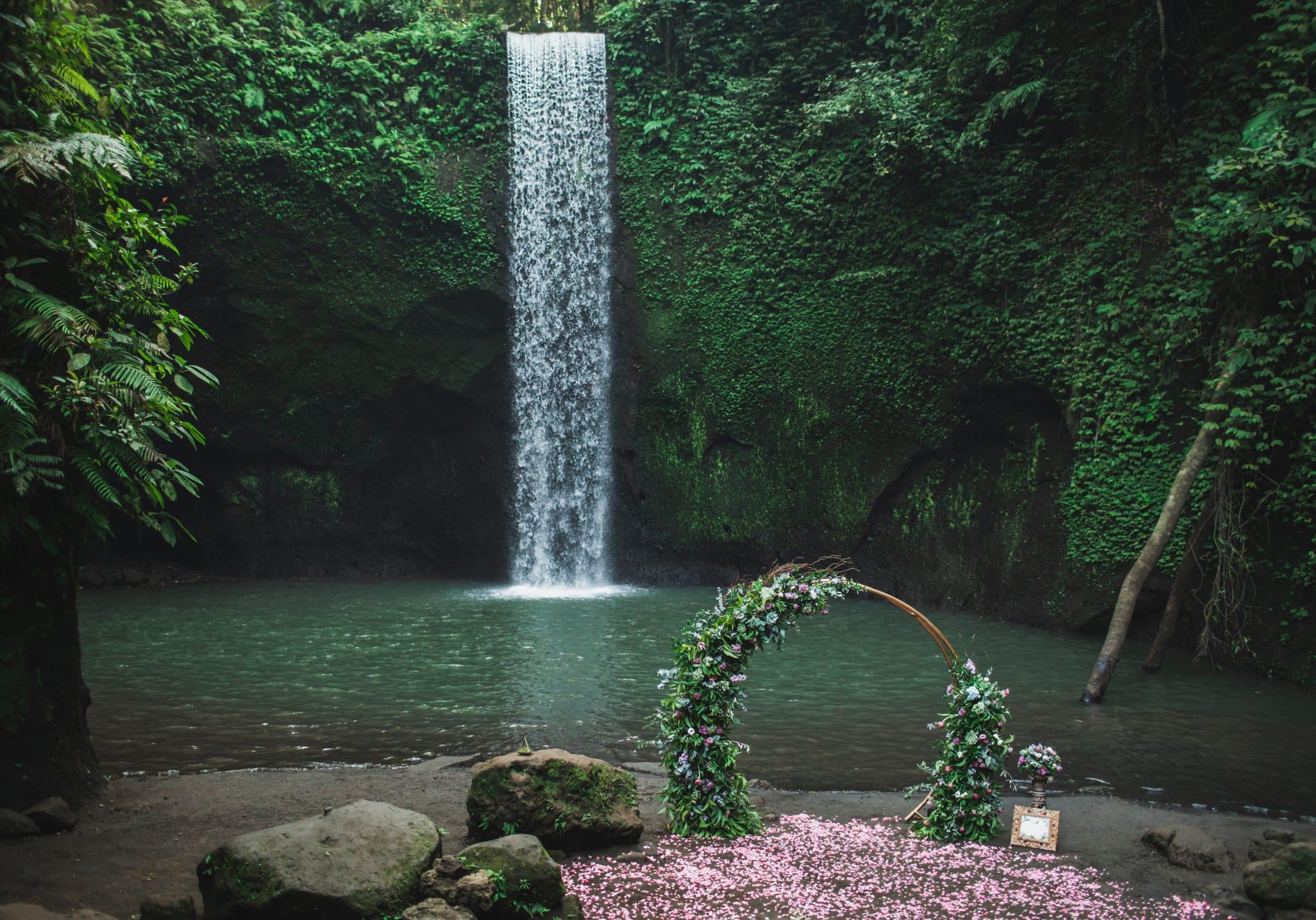 Round bronze wedding arch decorated with pink roses and greens. Unusual location for ceremony near small waterfall in jungle. Tibumana, Bali, Ubud.