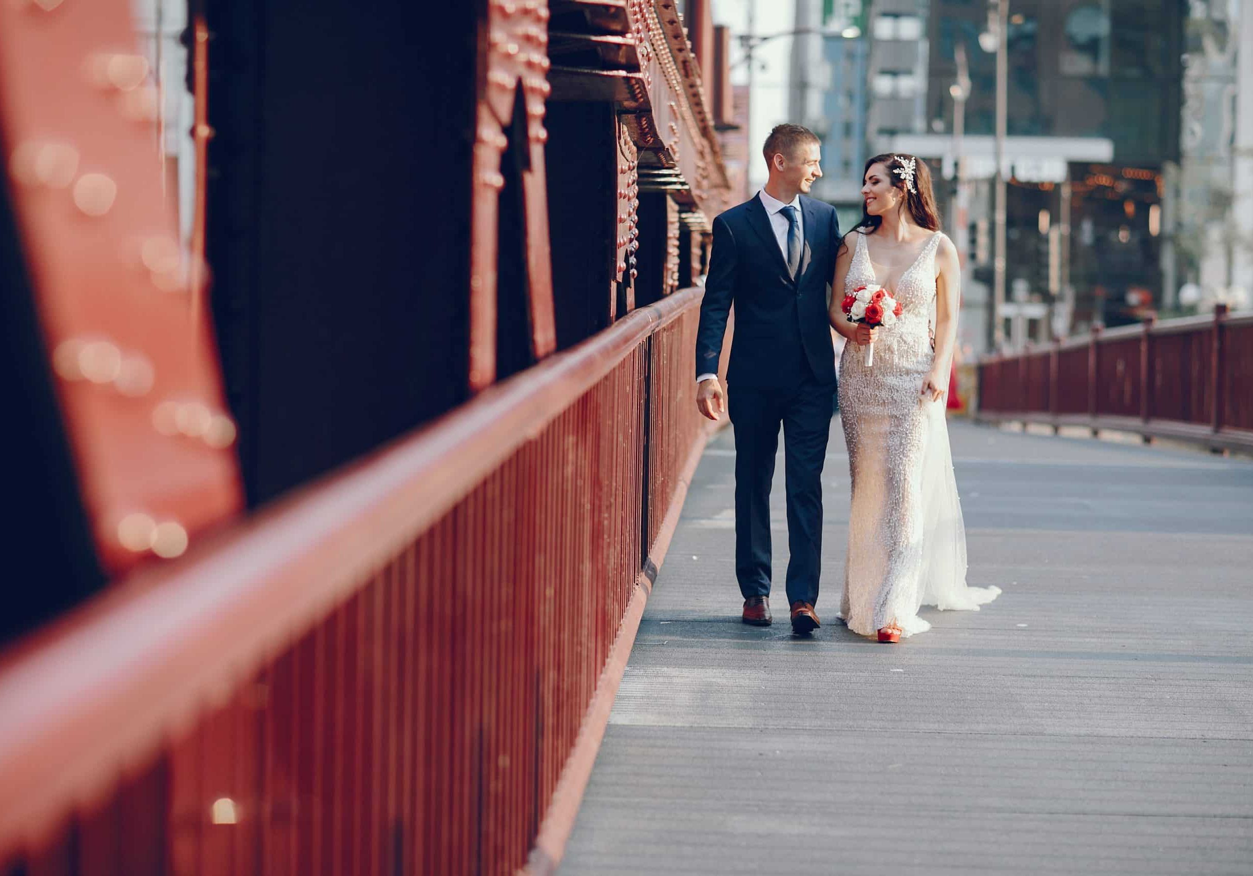 Elegant wedding couple walking in a summer city near brige. Man in a blue suit. Woman in a white elegant dress with bouquet of roses